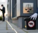 Technicien de dératisation posant une boîte d’appât noire scellée au pied d’un immeuble moderne, mains gantées en gros plan; inspecteur municipal flou, ambiance de conformité au cadre légal.