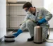 Technicien de dératisation et désinfection en combinaison gris clair, gants bleus et masque FFP2, agenouillé, place un boîtier d’appâtage scellé près d’une plinthe, pulvérisateur inox à tuyau jaune, salle technique propre.