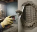 Reboucher un trou dans une cave ou un garage : prévention des intrusions; main gantée appliquant du mortier avec une truelle pour combler un trou au ras du sol dans un mur en béton, grille métallique anti-rongeurs partiellement insérée, outils flous en arrière-plan.
